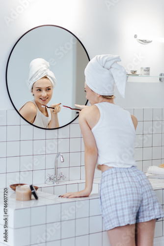 Blonde with a white towel on her head brushes her teeth in front of a mirror in a white bathroom.