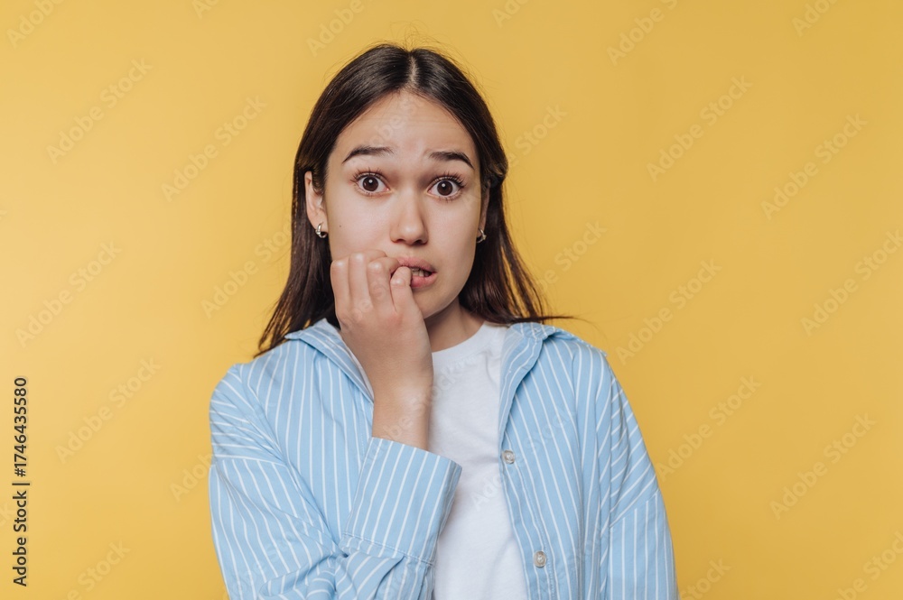 Obraz premium Young woman biting nails, looking anxious against yellow background.