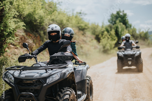 Photos A group of friends drives all-terrain quad bikes on an off-road forest path, showcasing adventure, teamwork, and outdoor exploration surrounded by lush greenery under a bright blue sky