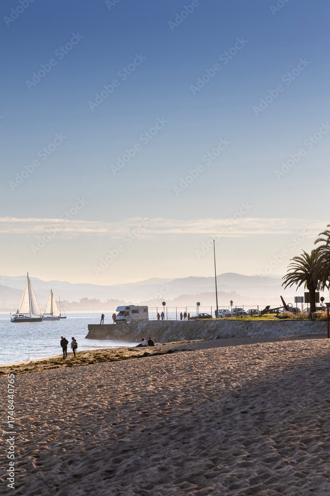 Obraz premium A scenic view of a tranquil beach in Santander, Spain, with sailboats on the water and mountains in the hazy distance. A concept of a peaceful seaside vacation.
