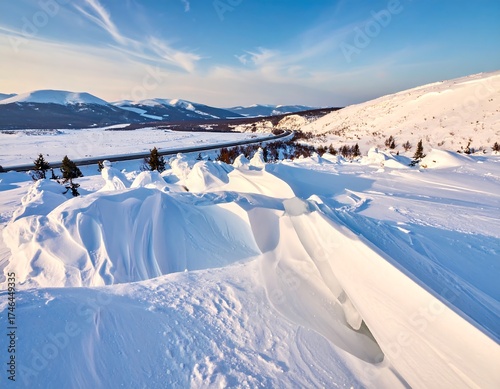 A snow-covered landscape with a road winding through mountains under a bright blue sky. Crisp, clean light highlights snow formations