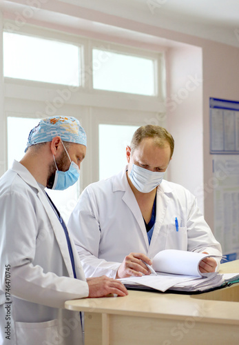 Two male doctors in protective medical masks discussing patient diagnosis and reviewing medical documents in a hospital. Concept of healthcare teamwork, professional medicine, and hospital routine.