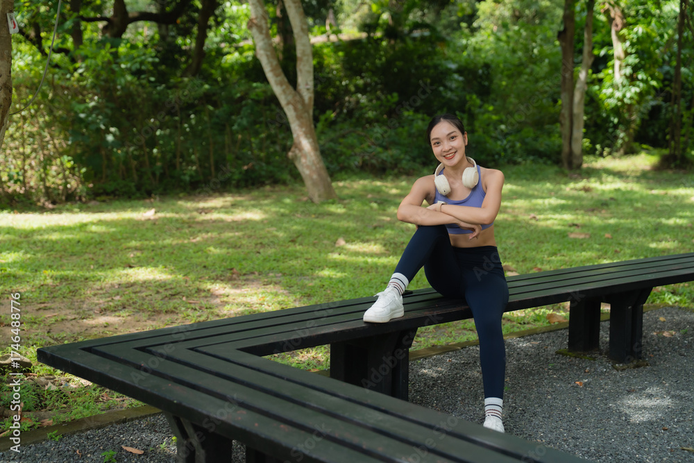Fototapeta premium Young Asian woman in sportswear sits on a park bench with wireless headphones around her neck, smiling at the camera while enjoying a restful moment outdoors after a workout