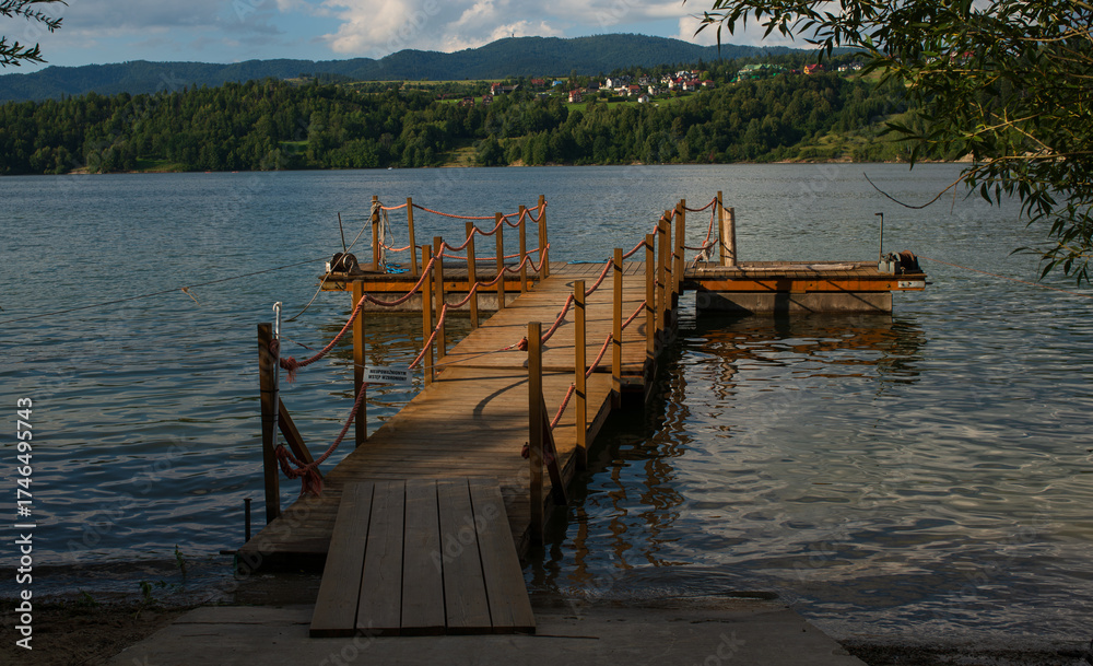 Fototapeta premium Wooden pier with orange ropes stretches into a quiet lake surrounded by forested hills and small village houses. Evening sunlight creates reflections and a sense of calm countryside beauty.