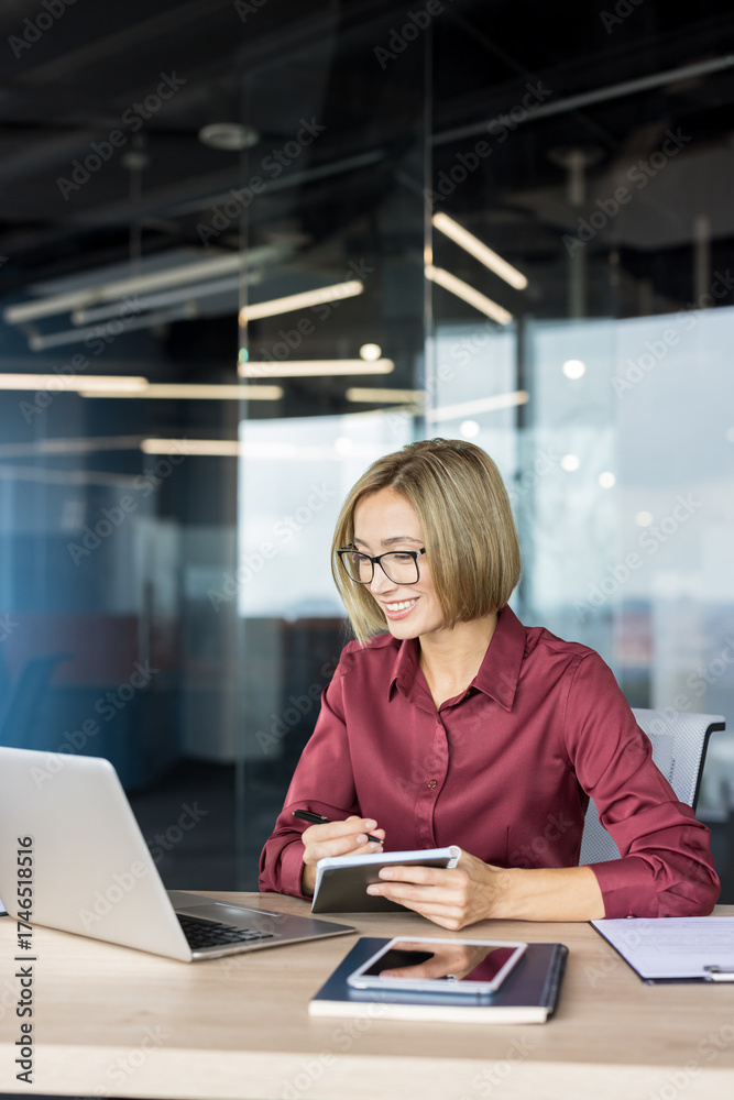 Obraz premium Smiling businesswoman in glasses taking notes while working on a laptop at her desk in a contemporary office, representing productivity and professional success
