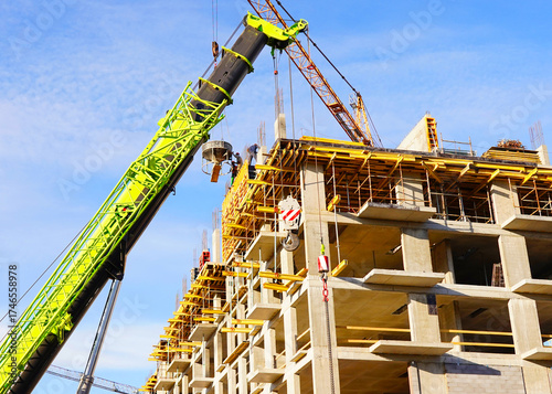 Building construction. Large green jib crane lifts materials to concrete structure. Construction workers in safety gear on scaffolding. Construction site with cranes hook. Formwork, pouring concrete.