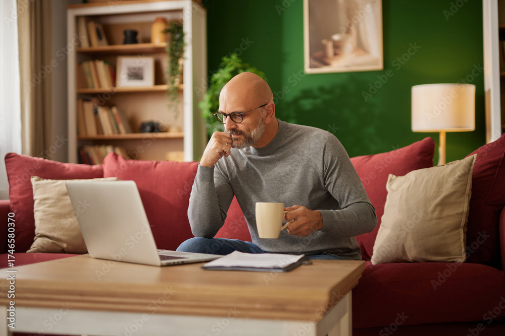 Fototapeta premium A man sits comfortably on a red couch, sipping coffee and analyzing financial documents on his laptop in a warm, inviting living room environment.