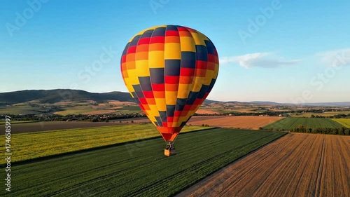 A vibrant hot air balloon ascends over lush green and golden fields under a clear blue sky with distant hills.