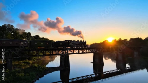 Steam train crossing a metal bridge over a calm river at sunrise or sunset, with smoke billowing and sun reflecting on the water.