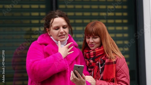 Two happy women standing outside, smiling, chatting, holding mobile phone, typing, browsing, blogging, swiping. People and technology. 