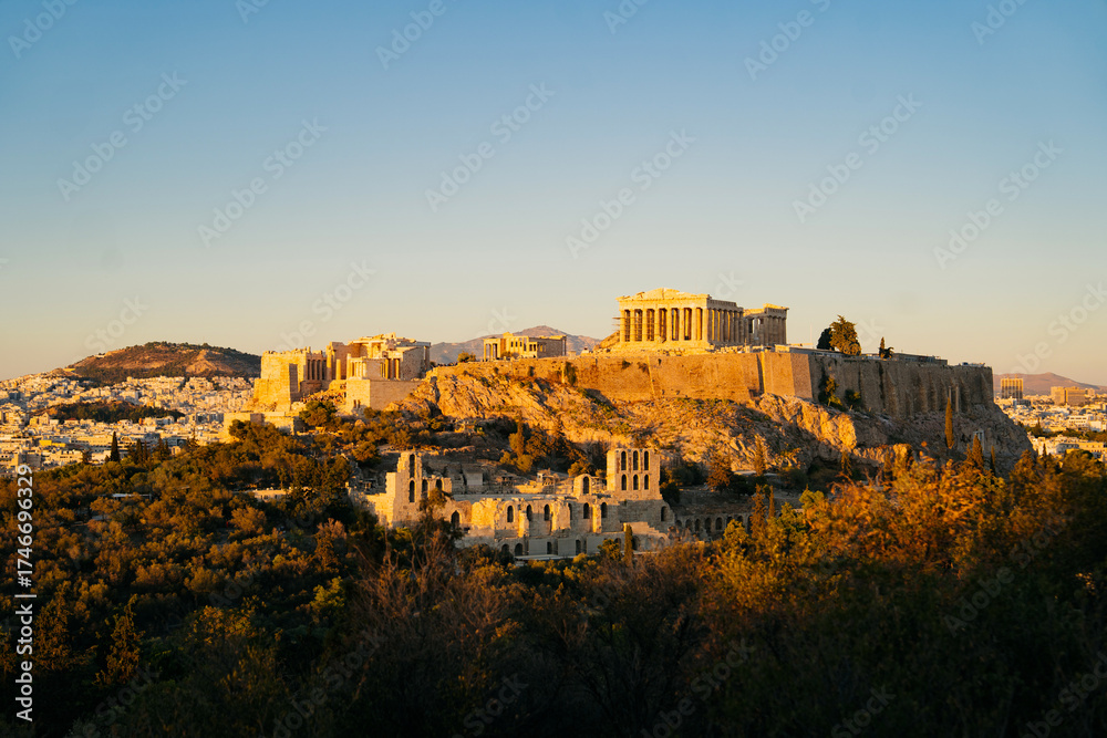 Fototapeta premium Acropolis of Athens during golden hour, with the Parthenon temple glowing above the city skyline. The soft evening light highlights the ancient ruins and the surrounding greenery, architecture