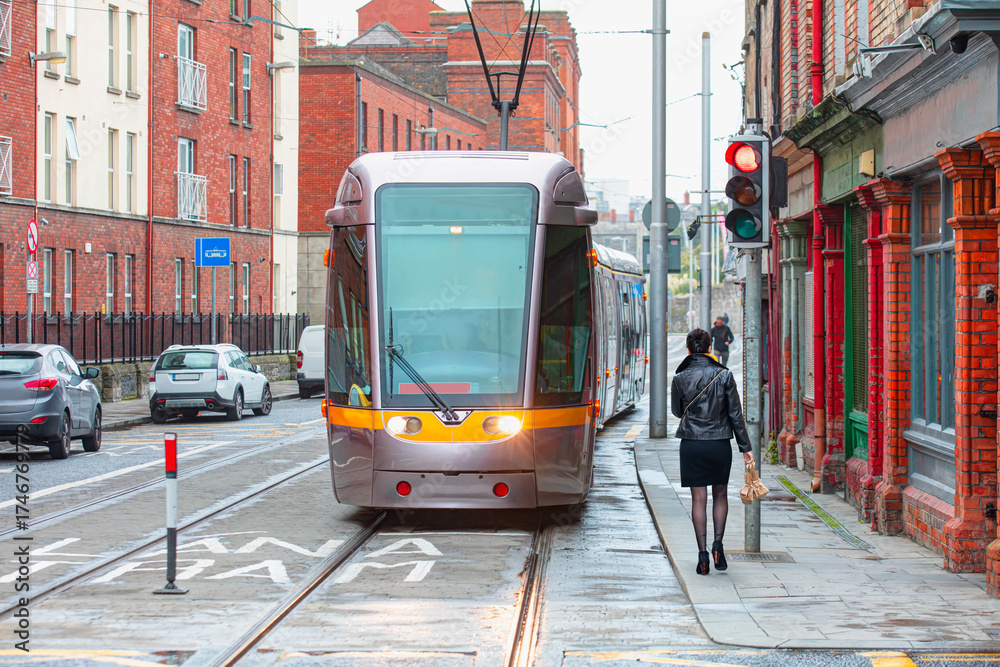 Fototapeta premium A well-dressed stylish woman is walking on the sidewalk next to traffic lights - Public tram on the street in Dublin, İreland