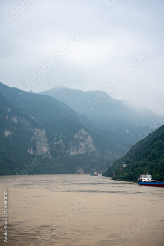 Yichang Three Gorges in China features a wide river flowing between immense, green mountains under a cloudy sky with boats navigating the water.