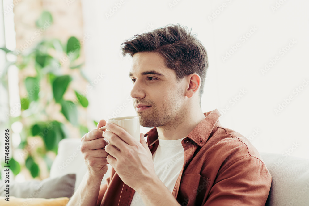 Fototapeta premium Young man enjoying morning coffee at home, sitting comfortably on a sofa in a bright modern interior