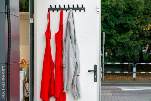 Interior of a fast food booth, open door with hanging red aprons and gray sweatshirt, view of street and greenery in background