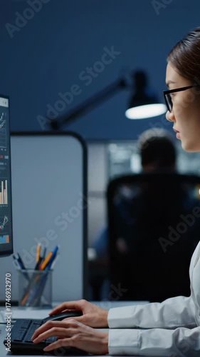 Focused young woman wearing glasses working on a computer in a modern office environment at night.