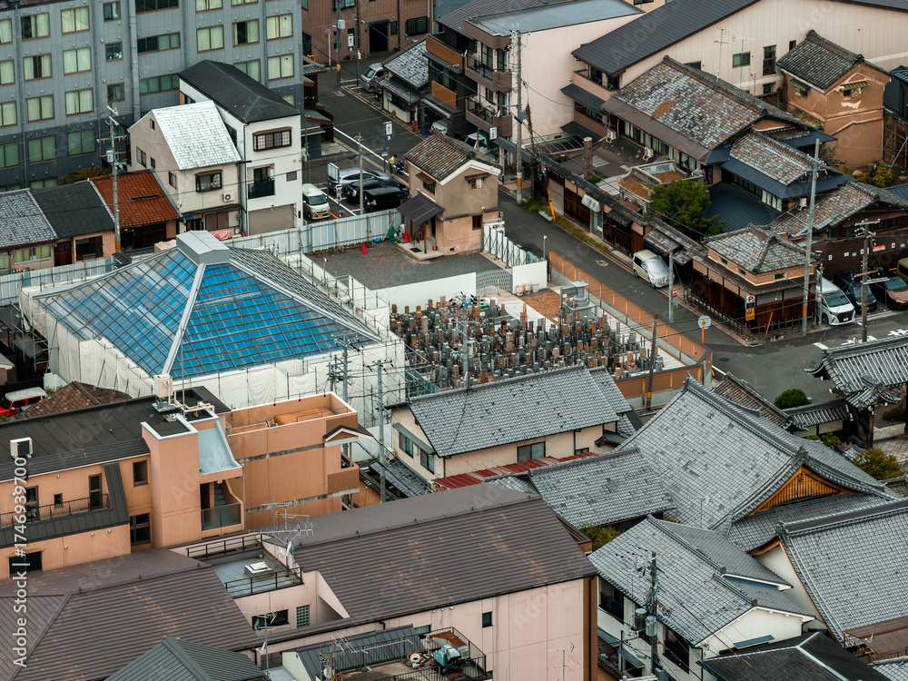 Fototapeta premium Aerial image of a cemetery with gravestones and a white fence in Kyoto, Japan, surrounded by traditional rooftops, modern buildings, and narrow streets.