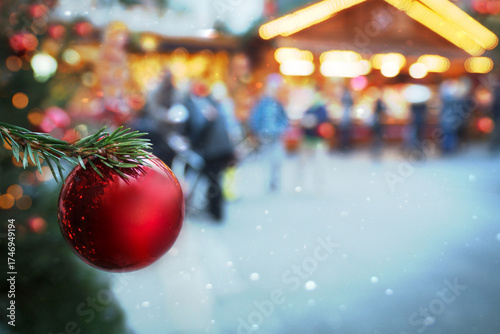 Rote Christbaumkugel, Weihnachtsschmuck, Tannenzweig auf Weihnachtsmarkt, Christkindlmarkt vor Verkaufsstand, Hütten, Schneefall, Schneeflocken, Lichtern, verschwommener Hintergrund im kalten Winter.