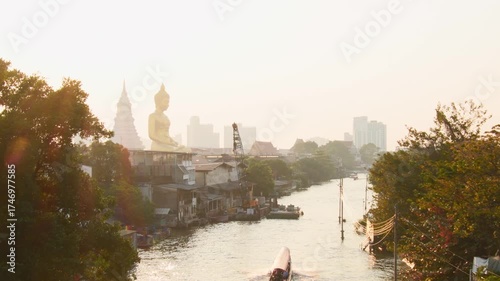 Golden Big Buddha statue at Wat Paknam Phasi Charoen temple in Bangkok seen above Khlong Phasi Charoen canal with traditional long-tail boats passing during sunset light