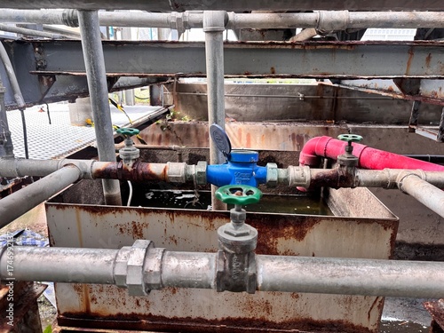 Close-up of a blue water meter and green valves on old, rusty industrial water pipes, illustrating aging utility infrastructure, plumbing issues, or water consumption measurement.