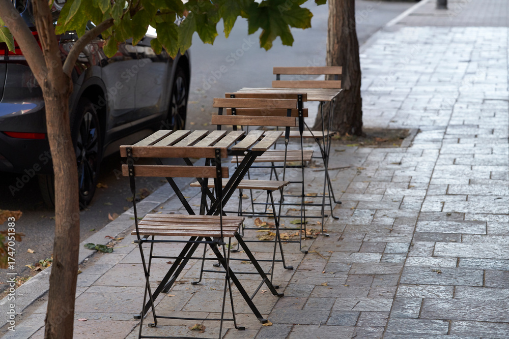 Obraz premium Tables and chairs at a summer cafe nestle among the trees on a city street. Fallen leaves litter the cobblestones. Autumn.