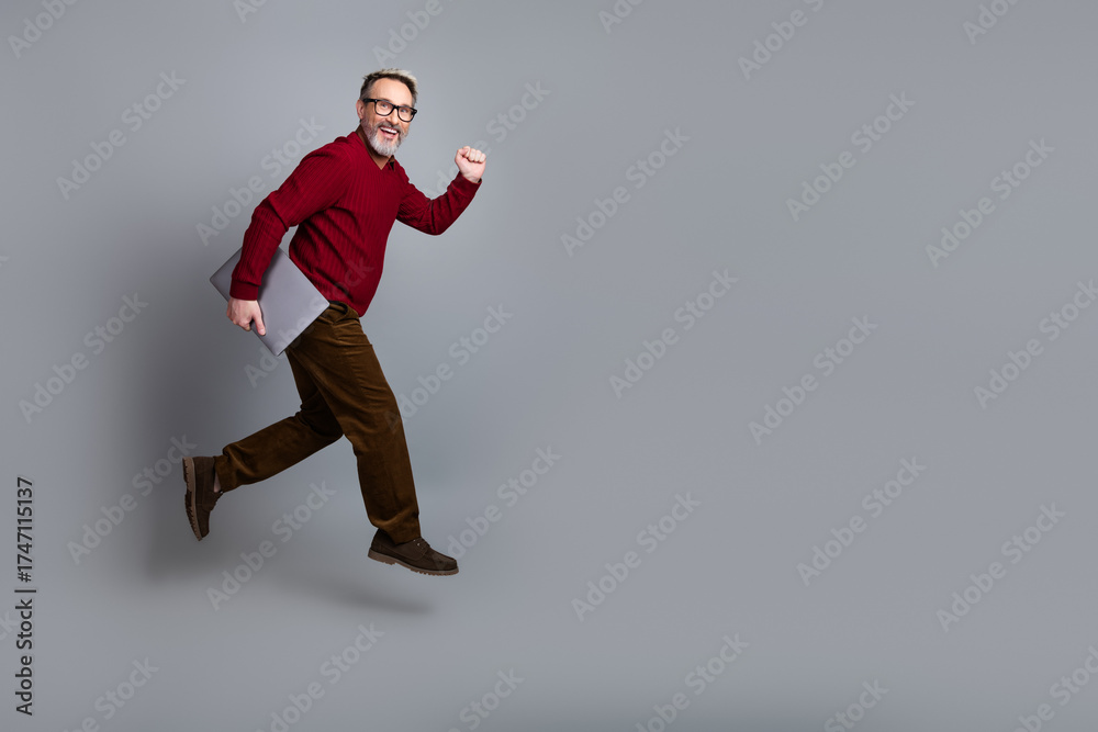 Fototapeta premium Handsome businessman holding a laptop in a red sweater mid-stride on a light gray background, exemplifying confidence and success