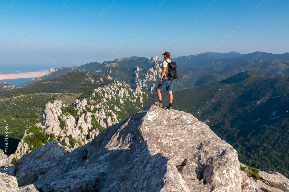 Fototapeta premium A lone hiker stands victoriously on a dramatic rocky peak in the Velebit mountains, overlooking the stunning Adriatic Sea coast in Kvarner Bay, Croatia on a summer day.