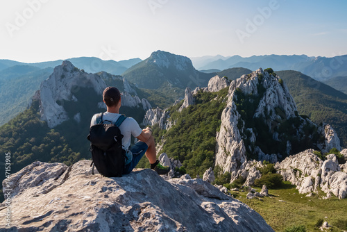 A male hiker with his backpack sits on a rocky peak in Croatia's Velebit mountains, contemplating the stunning and serene panorama of the majestic Dinaric Alps.