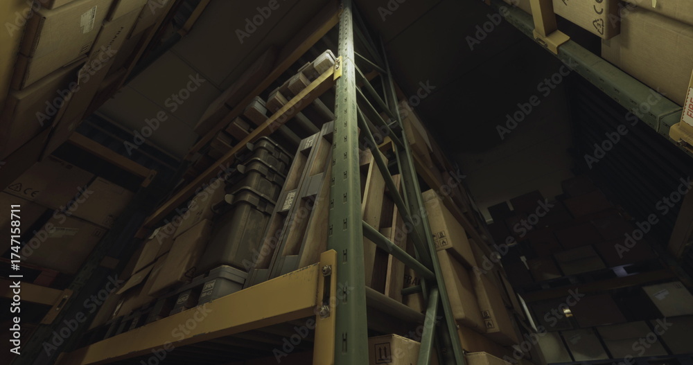 Fototapeta premium Tall shelves stacked with boxes fill a dimly lit storage facility. The structures create a maze like appearance, while shadows loom in the corners of the room.