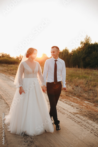 Bride and groom walking along a country road at sunset. The bride wears a beautiful white dress, the groom in a classic suit. Romantic wedding photo shoot outdoors with warm light and genuine emotions