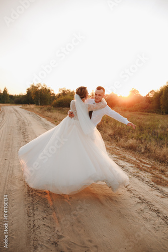Groom twirls the bride on a sandy road at sunset. Wedding photo shoot in nature capturing motion, joy, and tenderness. Romantic atmosphere with warm evening sunlight and true love.