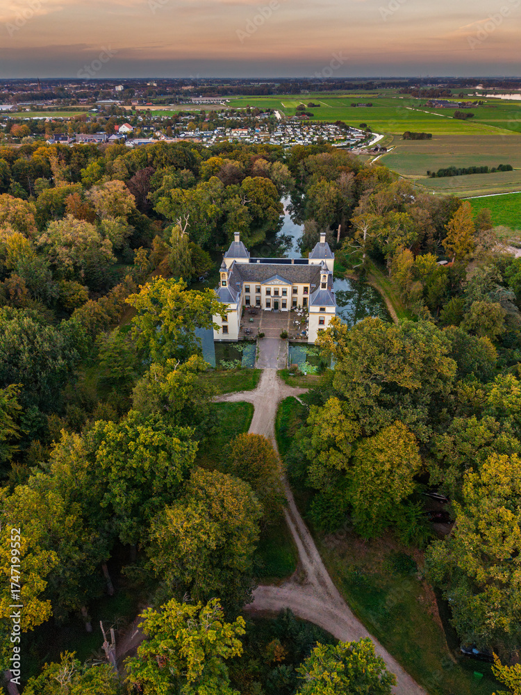 Obraz premium Aerial view of historic castle symmetrical twin towers and central hall framed by forest, moats, and tree-lined path, with open fields and residential zone beyond under warm sunrise sky.