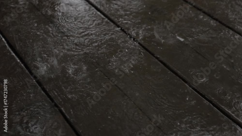 A soothing slow-motion video showing raindrops splashing on a wet wooden deck, forming ripples and reflections. The close-up view captures the calm rhythm and texture of rainfall in detail.