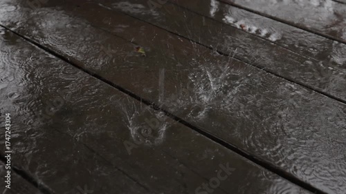 A soothing slow-motion video showing raindrops splashing on a wet wooden deck, forming ripples and reflections. The close-up view captures the calm rhythm and texture of rainfall in detail.