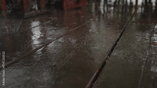 A soothing slow-motion video showing raindrops splashing on a wet wooden deck, forming ripples and reflections. The close-up view captures the calm rhythm and texture of rainfall in detail.
