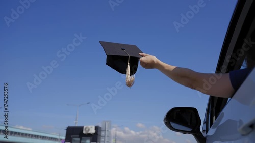 A Caucasian woman waves her graduation cap while extending her arms out of a car window.