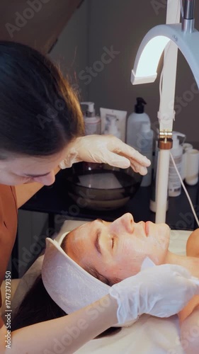 Cosmetologist in a beauty salon performs a skin cleansing procedure on a patient. The cosmetologist applies a mask to the woman's face to eliminate pigmentation and skin defects after acne.