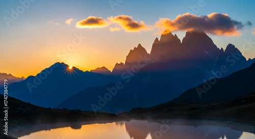 Majestic mountain peaks are silhouetted against a vibrant sunrise, with golden light illuminating the clouds and reflecting on the tranquil lake below