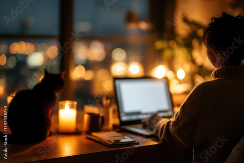 Person working on laptop at night with cat nearby, surrounded by warm candlelight and city view