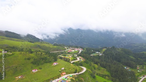 A beautiful aerial view of a remote mountain village in Flesha, Ainaro, Timor-Leste. A winding road crosses the lush green hills, with low clouds blanketing the distant peaks