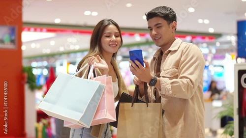 Happy Asian family couple using mobile phone taking selfie together while shopping at department store on holiday vacation. People enjoy urban lifestyle shopping, buying and spending at shopping mall.