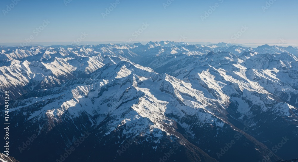 Fototapeta premium Aerial view of snow-capped mountain range under a clear blue sky, showcasing rugged peaks and valleys