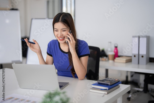 Quadro em tela Asian businesswoman happily discussing work on phone while using laptop at moder