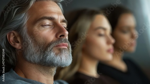 Close-up of a peaceful middle-aged man with gray hair and beard meditating with eyes closed alongside two blurred women also meditating indoors