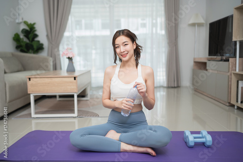 Young woman stretches in a modern living room while sitting on a yoga mat with dumbbells and a water bottle, promoting health and wellness lifestyle Free Download Young woman stretches in a modern living room while sitting on a yoga mat with dumbbells and a water bottle, promoting health and wellness lifestyle - GFXLoad