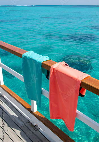 Turquoise and coral wetsuits hanging from a wooden railing overlooking a turquoise ocean. Vacationer's wetsuits drying on a boat deck in a tropical destination.