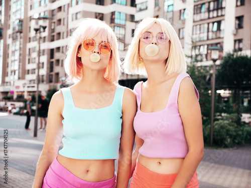 Two young beautiful smiling hipster female in trendy summer clothes. Sexy carefree women posing in the street in wigs. Positive funny models having fun at sunset. Blowing bubble with chewing gum