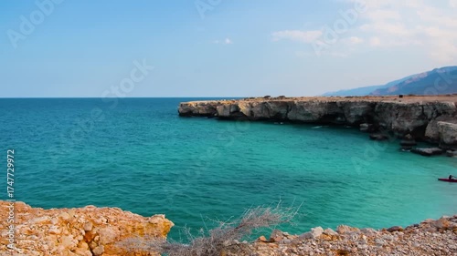 A view of the turquoise waters of the Arabian Sea meeting the rugged, rocky cliffs of the Omani coastline. A lone red kayak is visible in the distance, adding a sense of adventure to the serene, natur