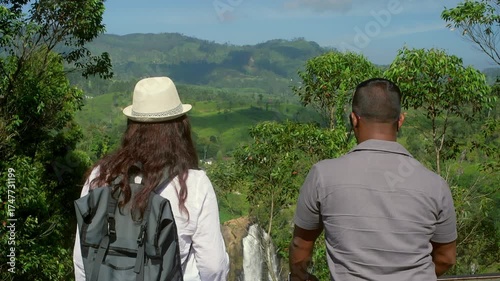Young travelers wearing backpacks and wide brimmed hats standing together, admiring cascading waterfall amid verdant tea plantation landscape in scenic Sri Lanka
