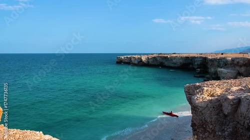 aerial view captures a lone kayaker navigating the stunning turquoise waters along the rugged, rocky coastline of Qalhat, Oman. The clear blue sky and dramatic cliffs create a serene and adventurous s